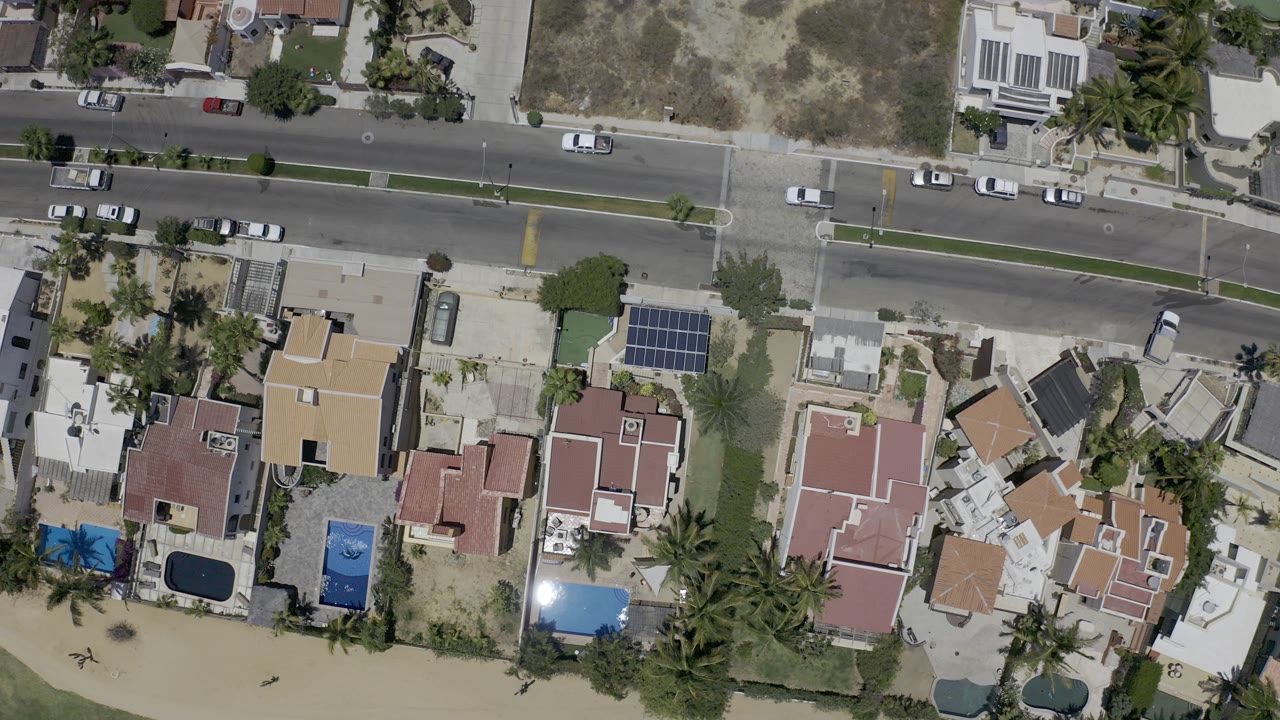 Overhead view of a coastal road, homes, pools, palm trees, and beach edge.
