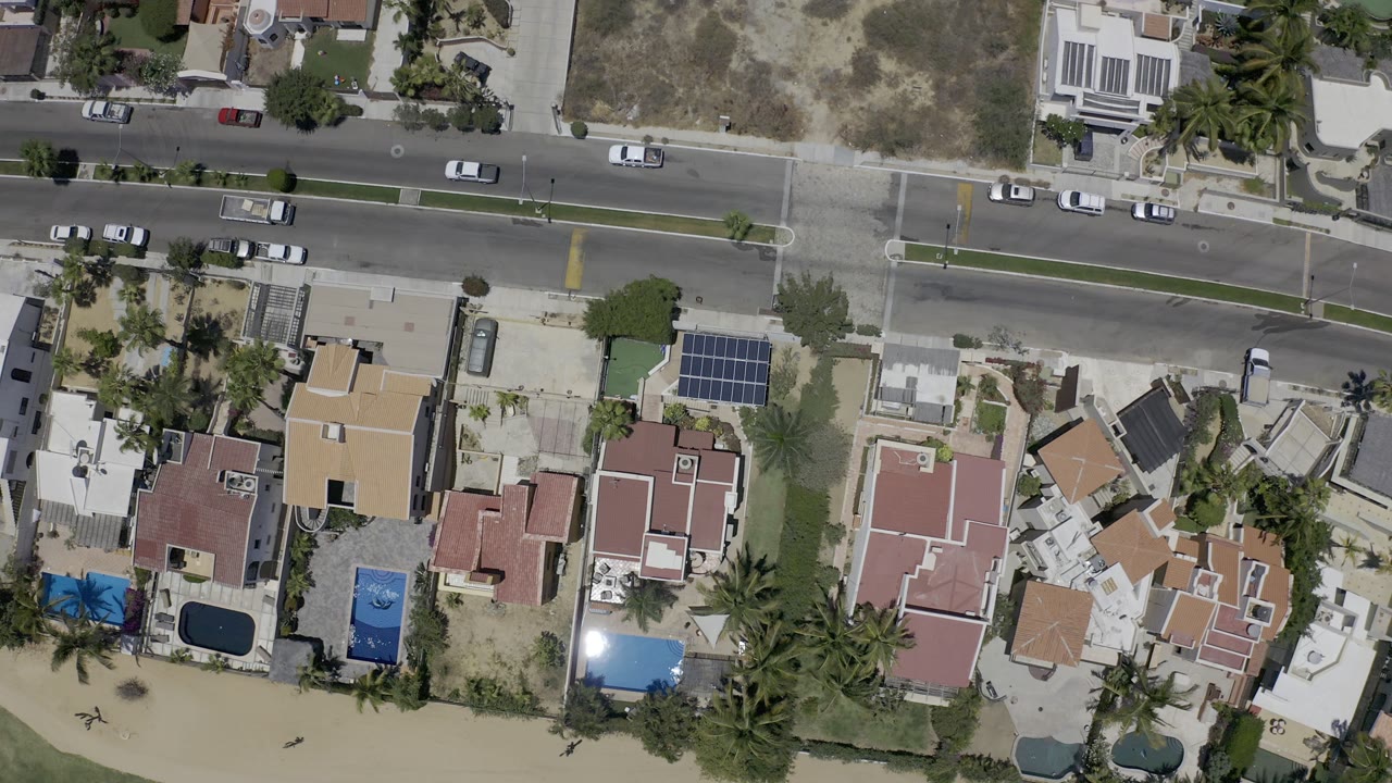 Overhead Los Cabos neighborhood view with the road centered near the top, compact homes and pools below it, sandy beach along the lower edge, and no visible people.