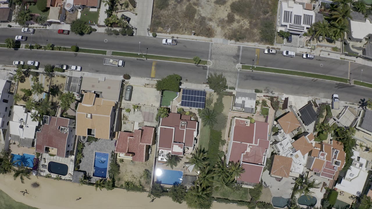 Second overhead frame in Los Cabos showing the same residential street, bright roofs, parked cars along the road, dry lots, palms, and no visible people.