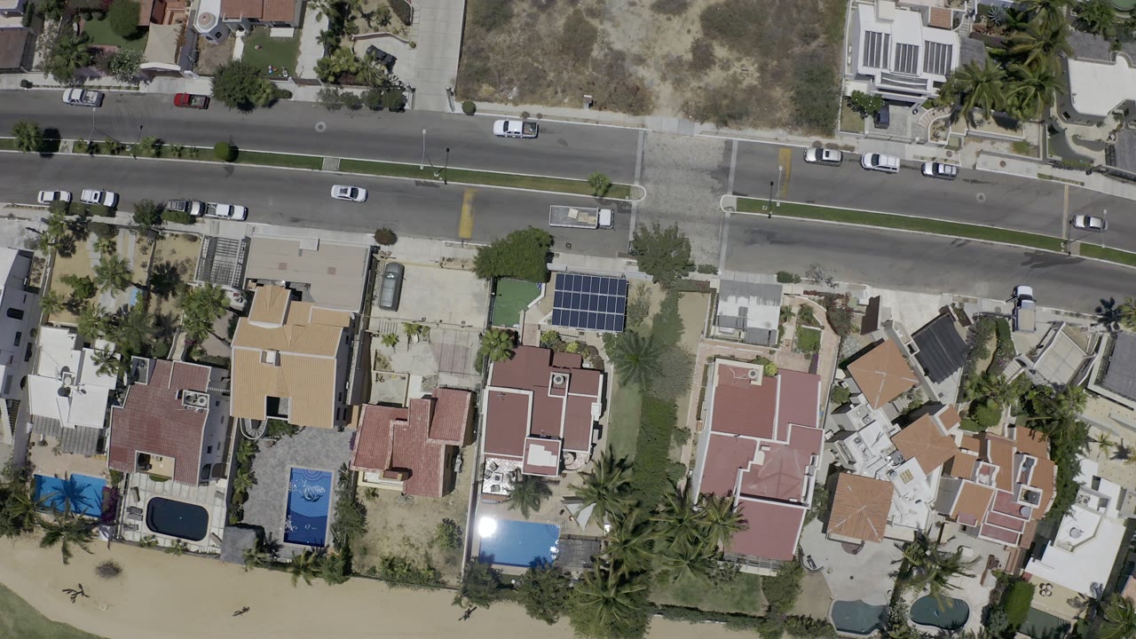 At 0:01, the aerial frame shows cars spaced along the Los Cabos road, with houses, a blue-roofed structure, palms, pale sand, and no visible people.