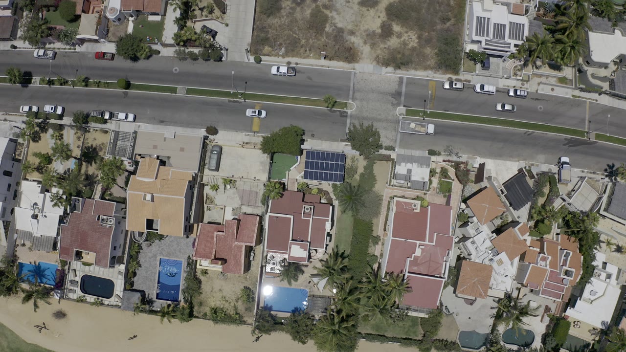 Overhead view of houses, pools, a road, parked cars, and beach sand.