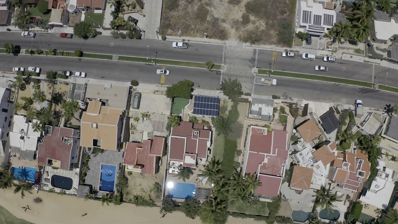 High overhead Los Cabos view with the road across the upper frame, villas and pools below, solar panels on one roof, palm trees near the beach edge, and no visible people.