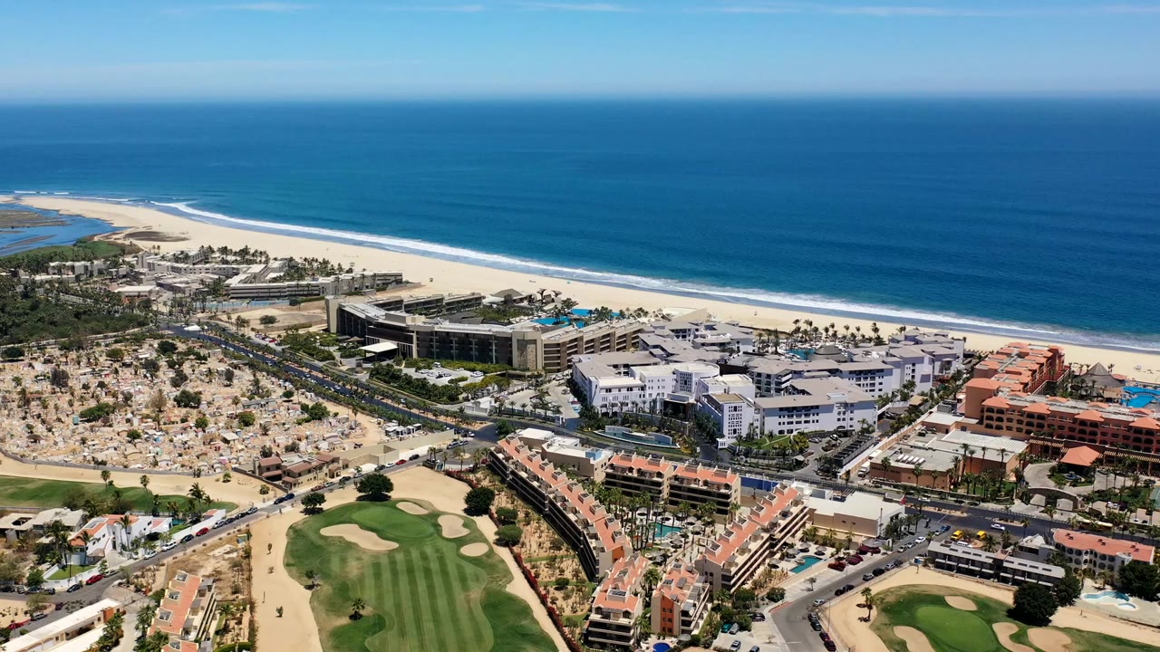 Aerial frame over Los Cabos, Mexico with the resort complex centered, golf course and road below, pale beach behind the buildings, and small distant beachgoers near the shoreline.