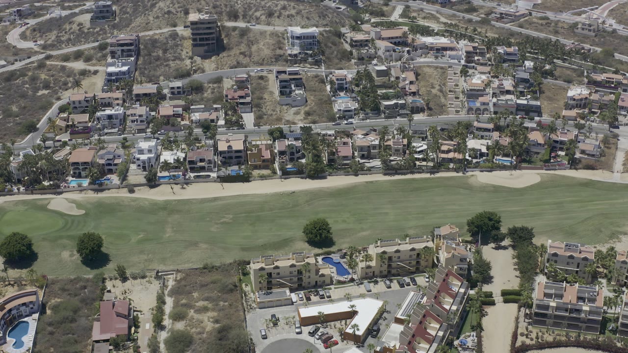 Aerial Los Cabos view with the green field centered, homes and planted trees across the middle, and dry hillside roads and larger buildings in the background, with no visible people.