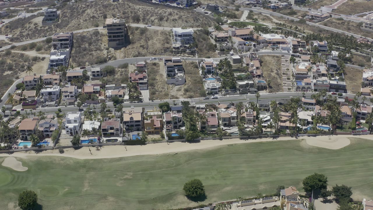 Aerial view in Los Cabos, Mexico, with fairway grass at the bottom, a line of homes and palms near the road, and dry hillside roads above, with no visible people.