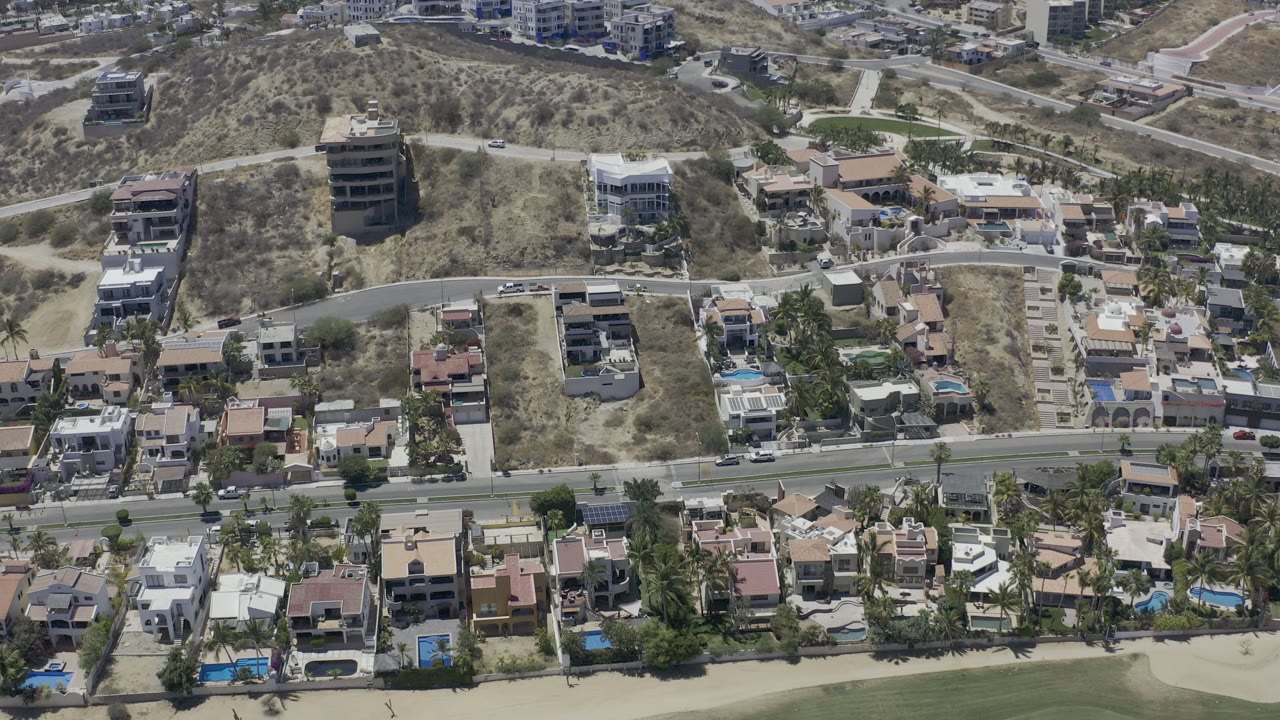 High aerial frame over Los Cabos showing hillside houses, a road across the lower frame, dry scrub slopes, small pools, and no visible people.