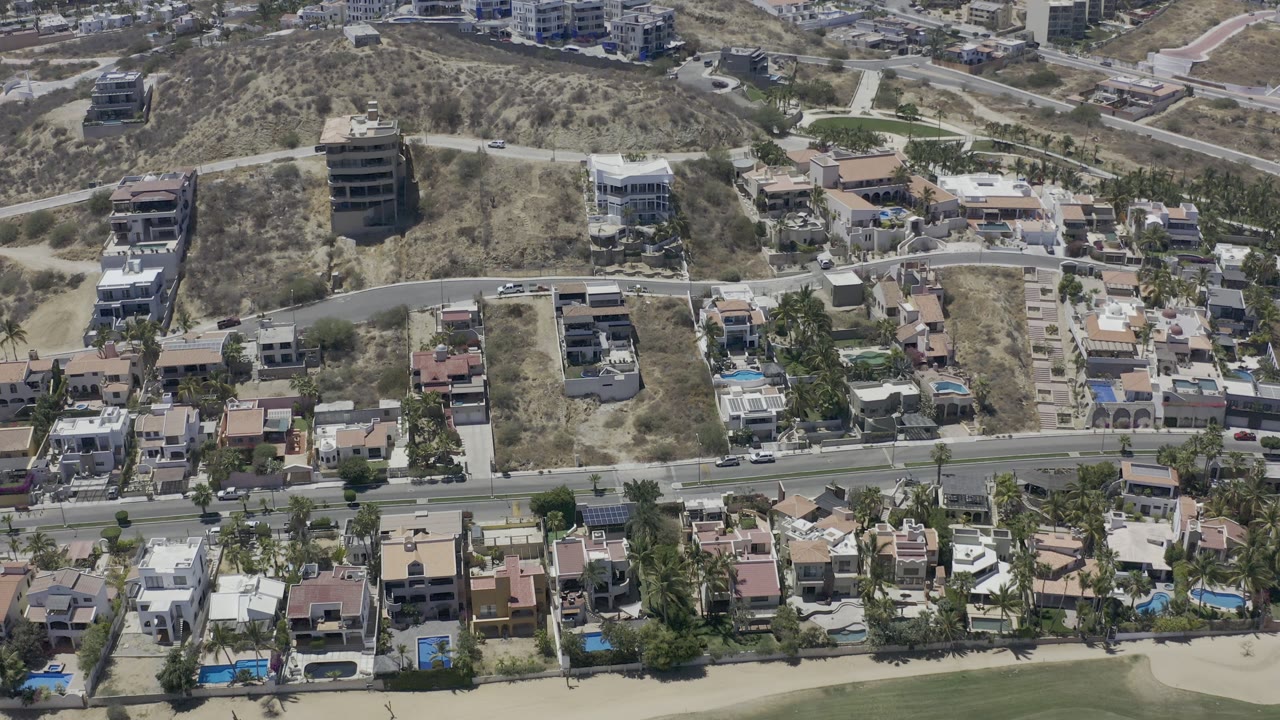 Aerial frame in Los Cabos with the main road lower in view, residential villas and palms below it, dry hillside lots above, and no visible people.