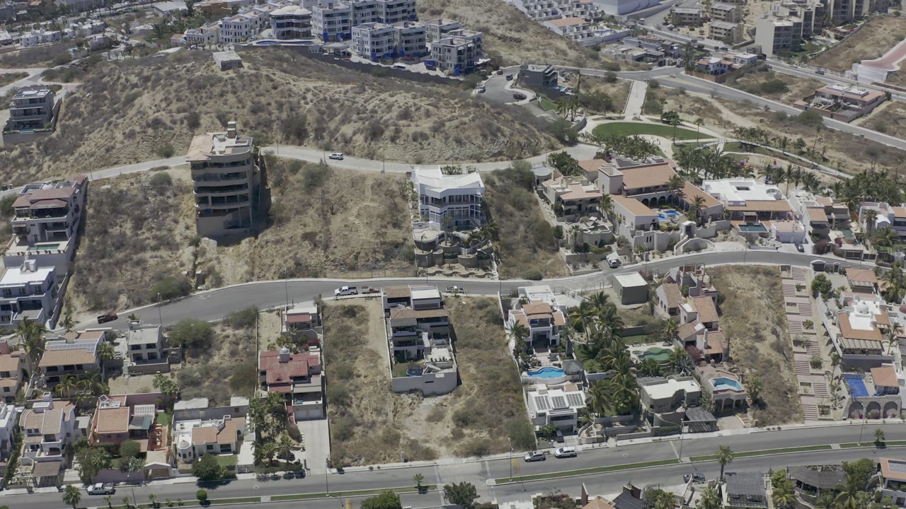 High aerial view in Los Cabos showing a brown hillside, terraced residential lots, white apartment buildings, roads, parked cars, and no visible people.