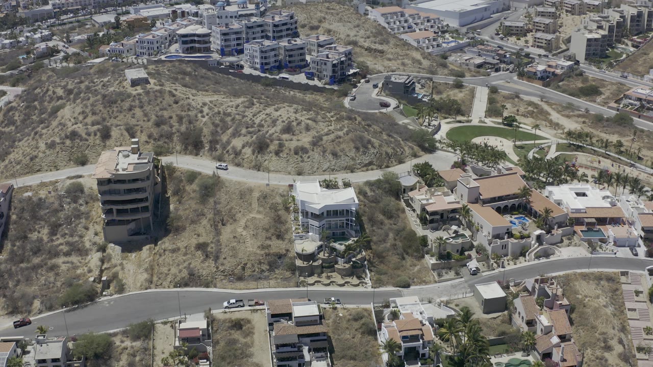 Aerial view in Los Cabos, Mexico, showing dry scrub hills, curving roads, modern homes, and distant buildings, with no visible people.