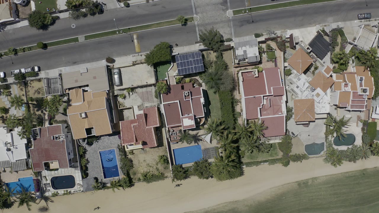 Top-down aerial view of coastal homes, pools, sand, and a road.