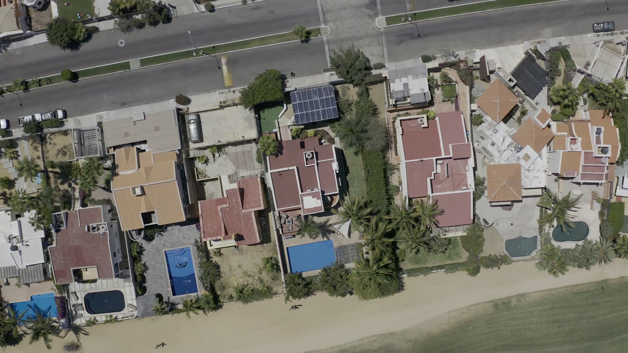 Overhead Los Cabos view centered on several private homes with pools, palm landscaping, solar panels, road lanes, and no visible people.