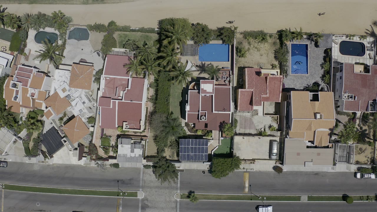 Overhead frame from Los Cabos, Mexico, showing red-roofed homes, blue pools, palm trees, a paved road, sandy shoreline, muted water, and no visible people.
