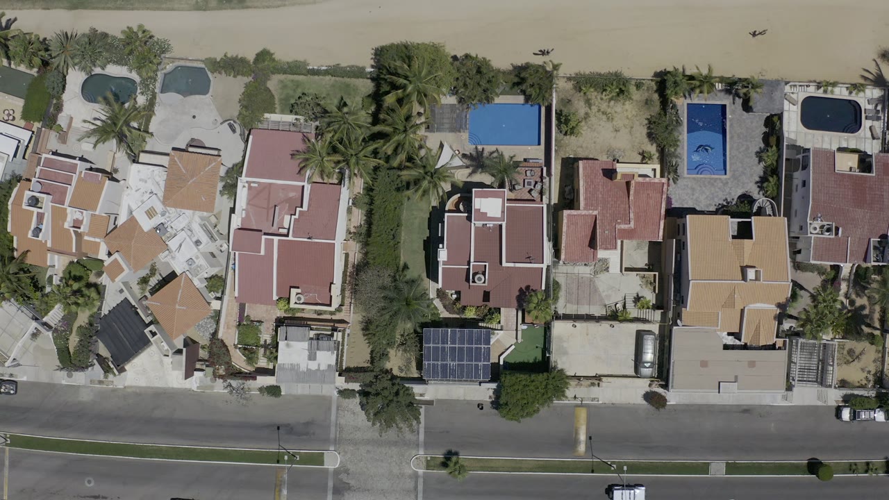 Straight-down Los Cabos aerial view with clustered red and tan roofs, rectangular blue pools, palms, a road across the lower frame, shoreline sand, and no visible people.