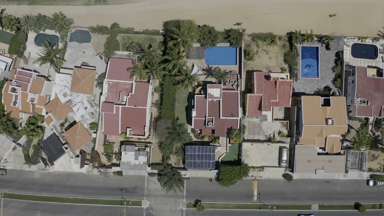 Aerial view of coastal homes, pools, parked cars, road, sand, and water.