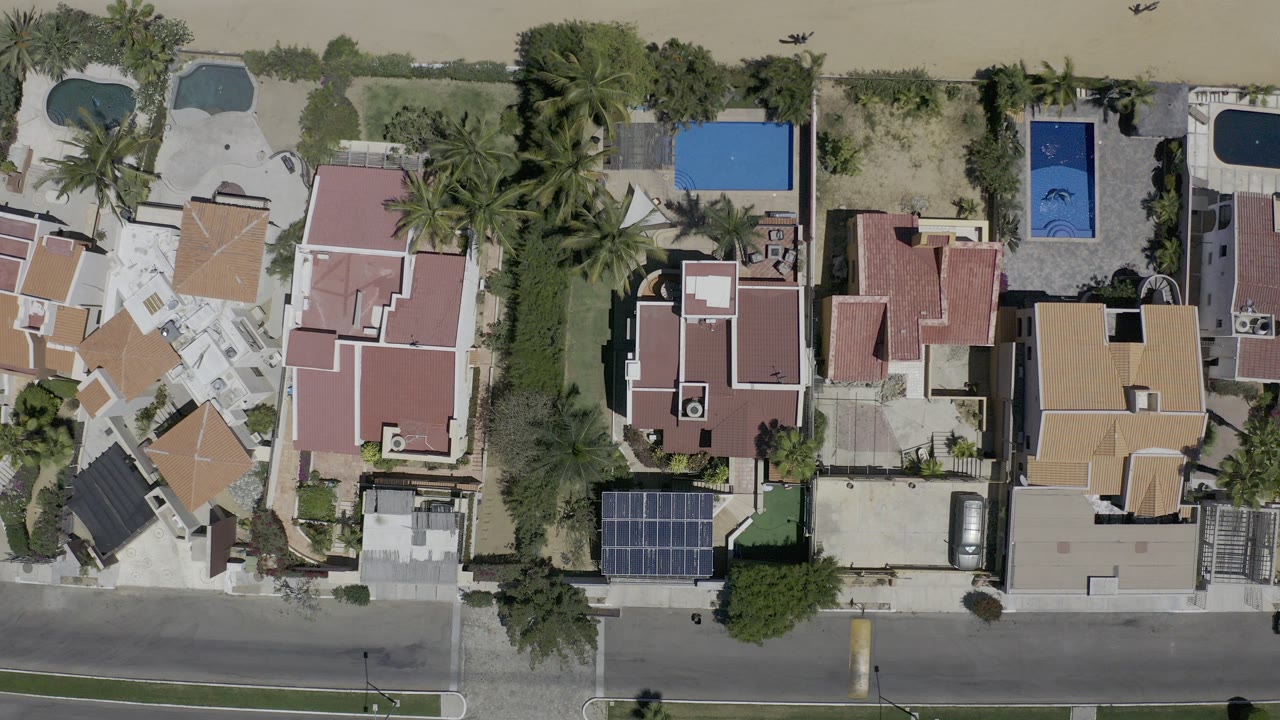 Overhead frame in Los Cabos, Mexico showing private homes, blue pools, palm trees, driveways, sandy ground, and no visible people.