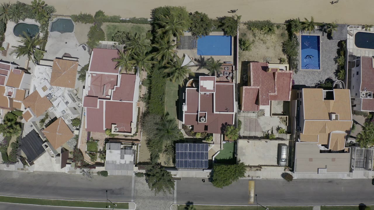 Aerial frame in Los Cabos, Mexico with rooflines, walled yards, multiple swimming pools, palms, a paved street, and no visible people.