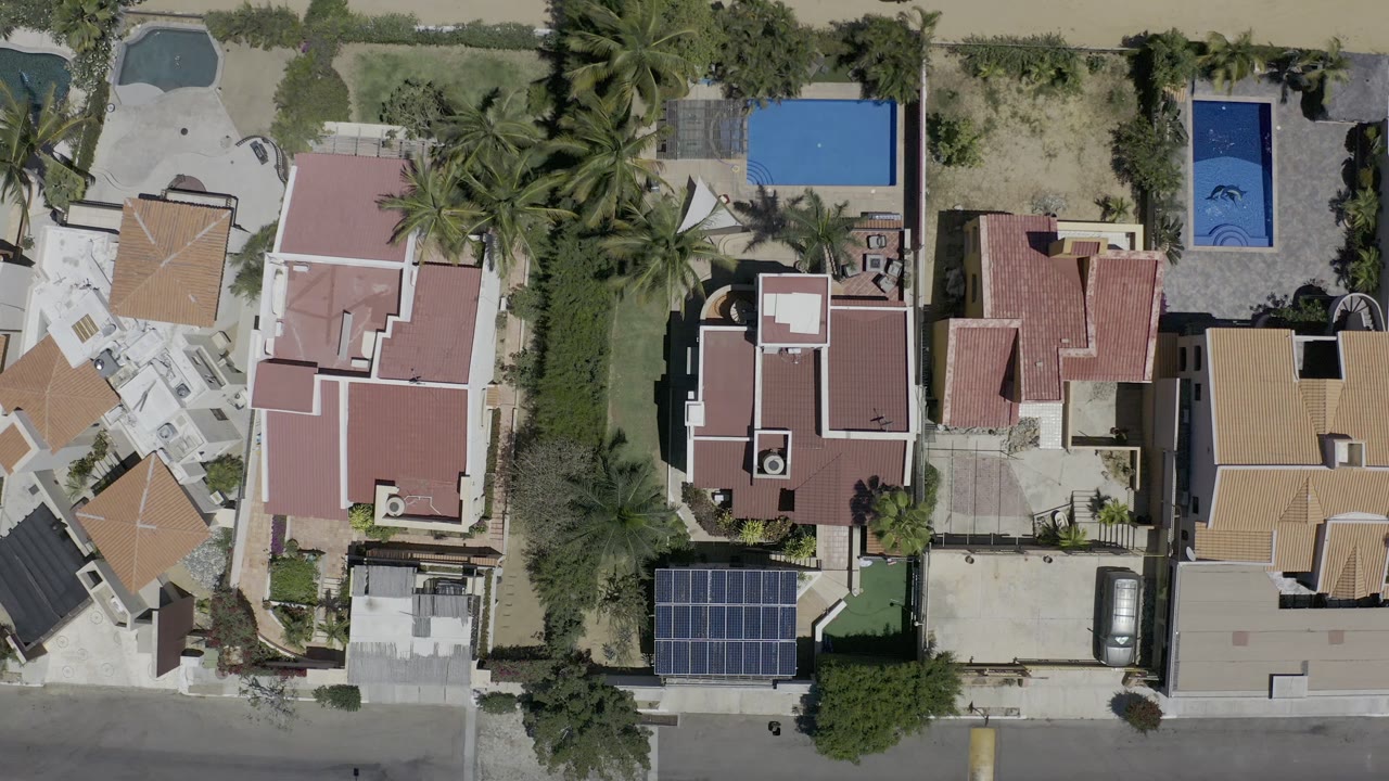 Top-down Los Cabos frame with multiple vacation rental roofs, a narrow palm-filled property line, blue pools near the top edge, solar panels near the street, and no visible people.