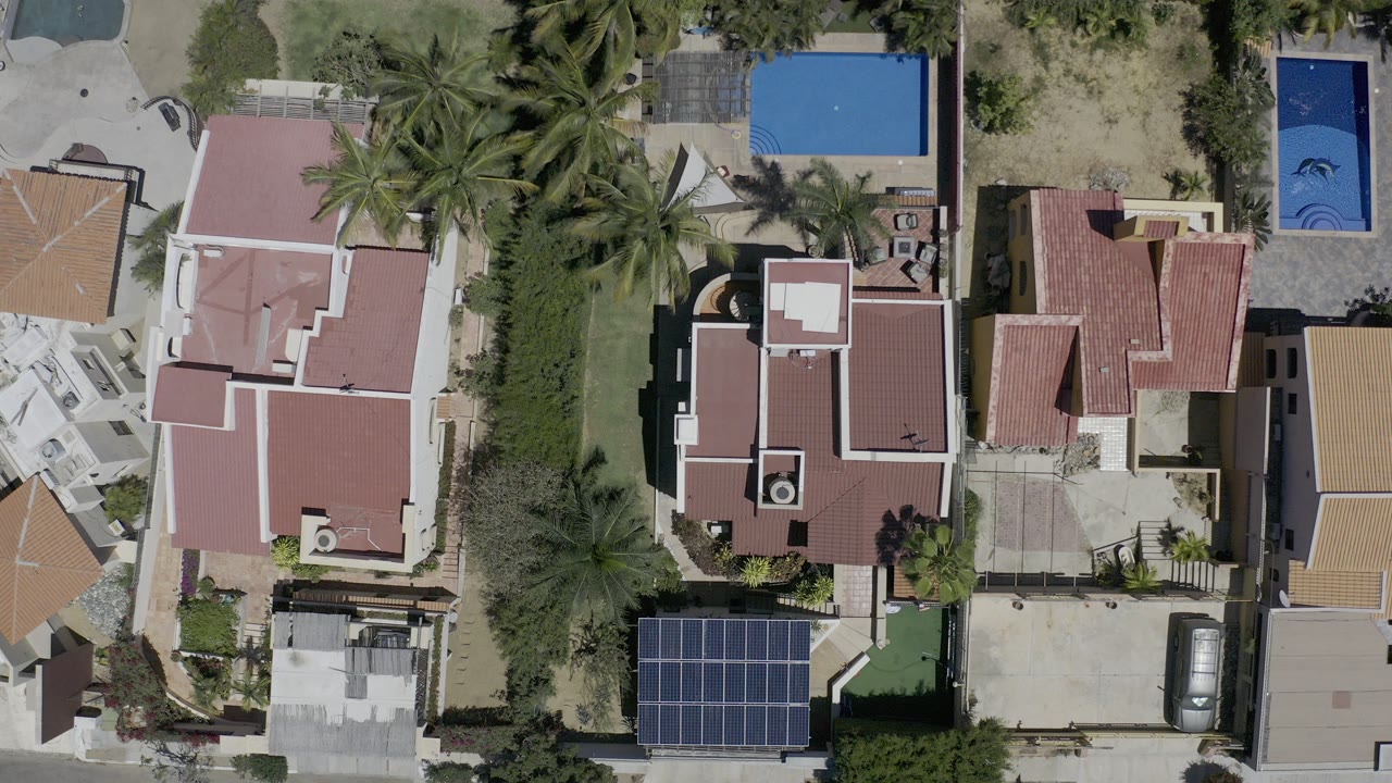 Overhead Los Cabos frame showing red-roofed homes, palm trees, blue pools, driveways, a solar panel array, parked cars, and no visible people at the start of the clip.