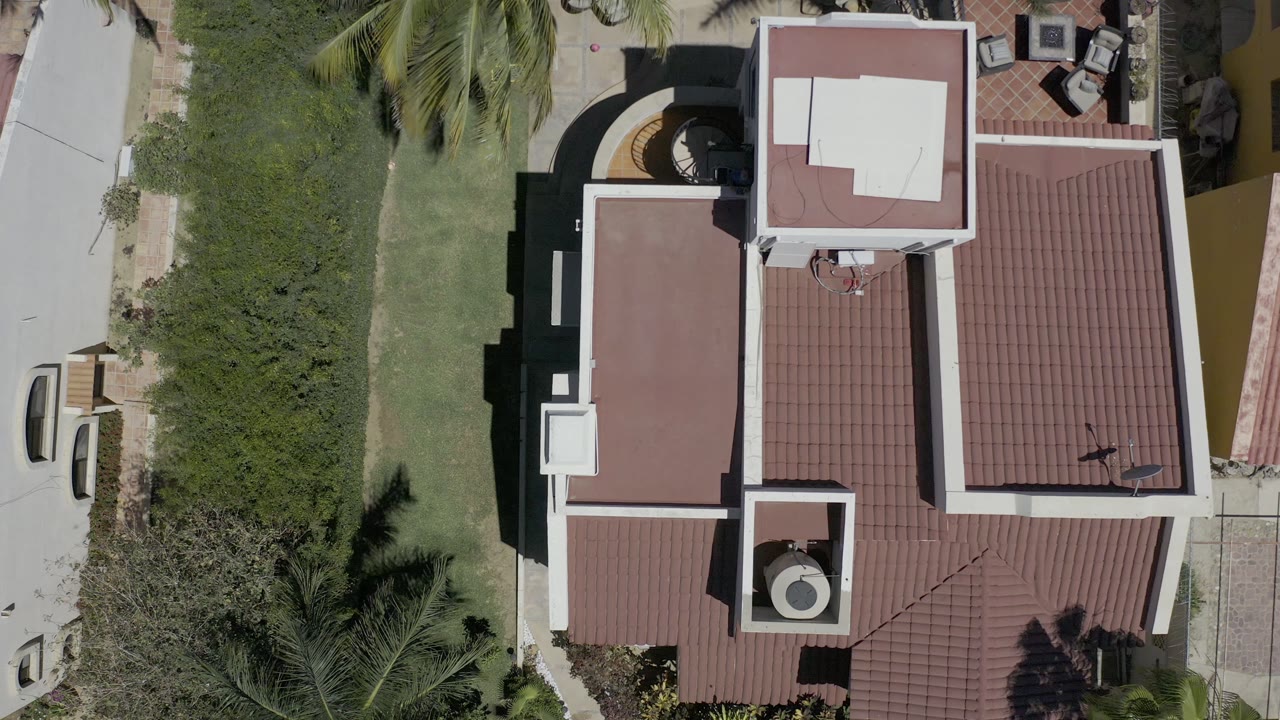 Top-down Los Cabos frame with the white building centered, red tile roof planes, green lawn to the left, paved access at the edge, and no visible people.