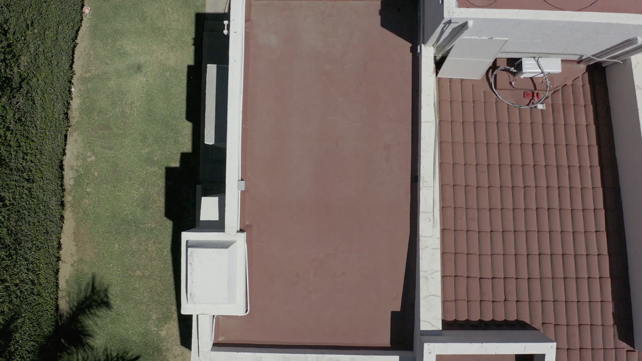 Top-down aerial frame in Los Cabos, Mexico, showing a red flat roof beside red barrel tiles, green lawn, hedges, white parapet walls, and no visible people.
