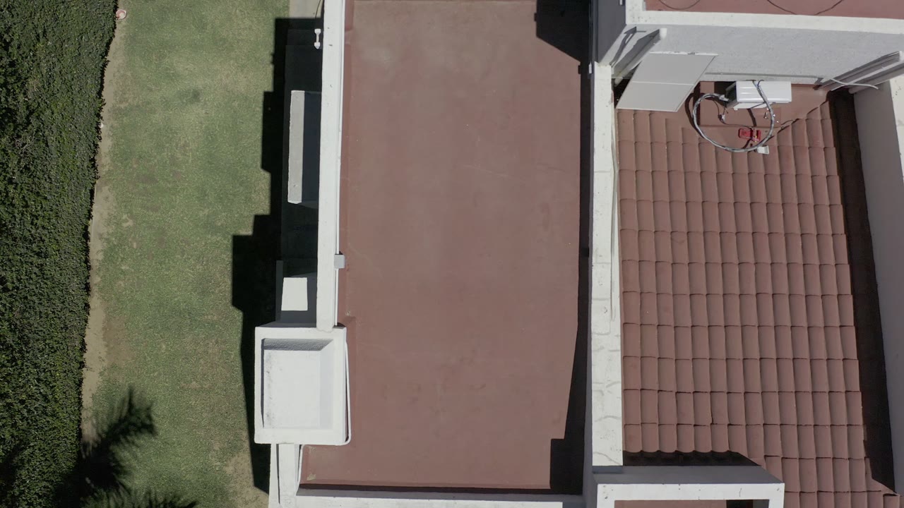 Aerial view over a Los Cabos rooftop, with the flat red roof centered, tile rows on the right, green lawn on the left, and no visible people.