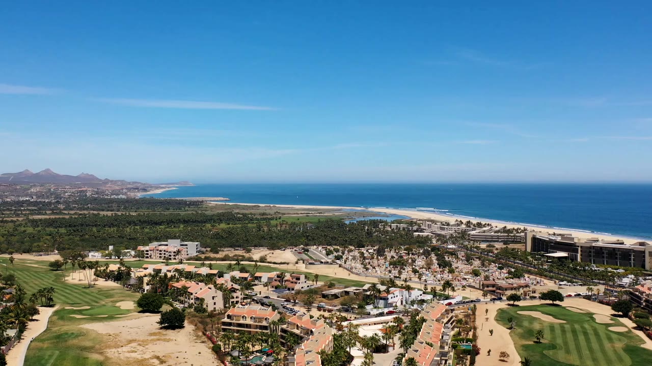 Los Cabos aerial view with terracotta-roof homes in the lower frame, green resort grounds, a long beach, distant mountains, and no visible people.