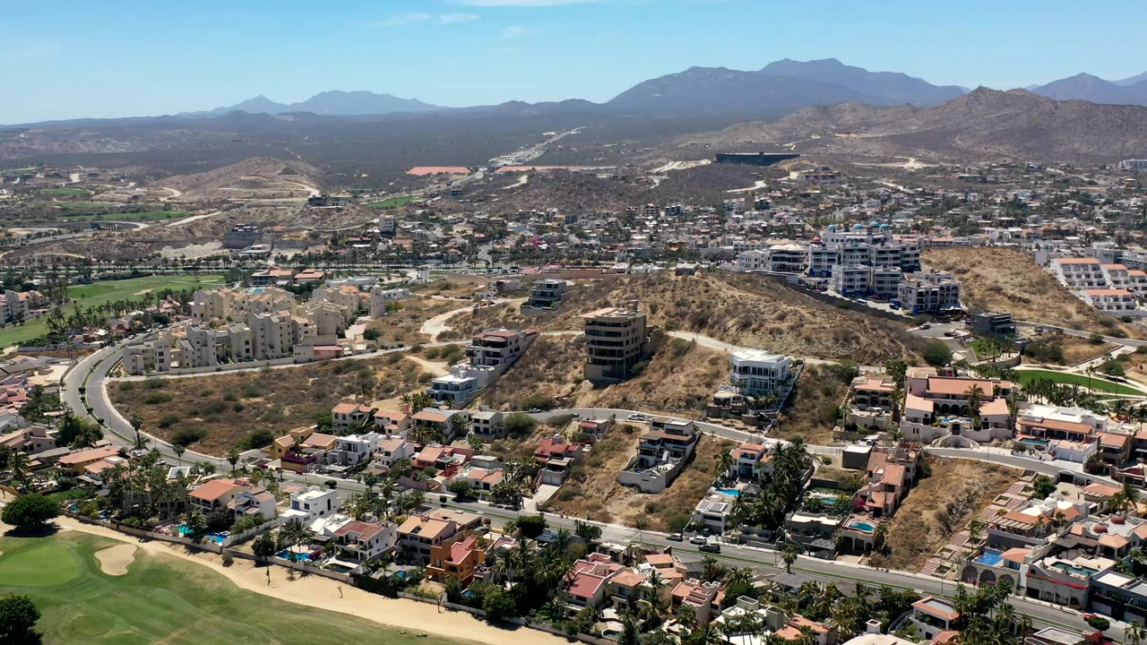 High aerial view over Los Cabos, Mexico, with hillside homes, curving roads, green grass at lower left, dry terrain, distant mountains, and no visible people.