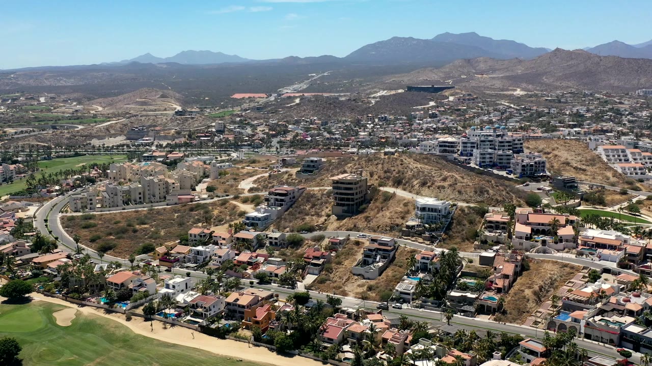Aerial frame over Los Cabos, Mexico, showing residential buildings spread across tan hills, a road network, small vehicles, mountain ridges, and no visible people.