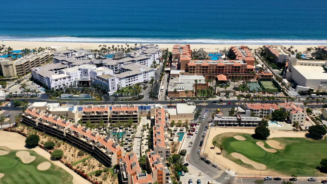 Opening aerial frame over Los Cabos, Mexico, with ocean surf beyond beachfront buildings, red-tile condo roofs, streets, pools, and a green golf course in the foreground, no visible people.