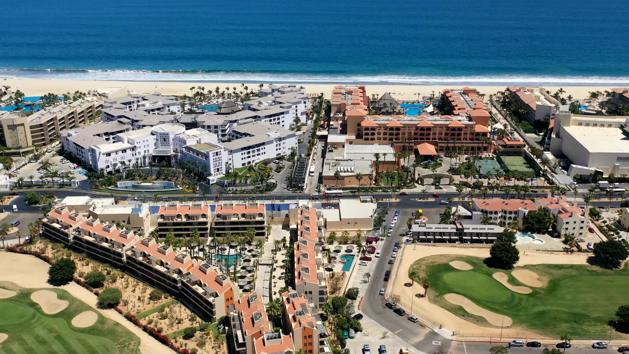 Aerial view in Los Cabos, Mexico, showing a white condo complex beside red-roofed buildings, a beachfront strip, blue ocean, and golf greens with sand traps below, no visible people.