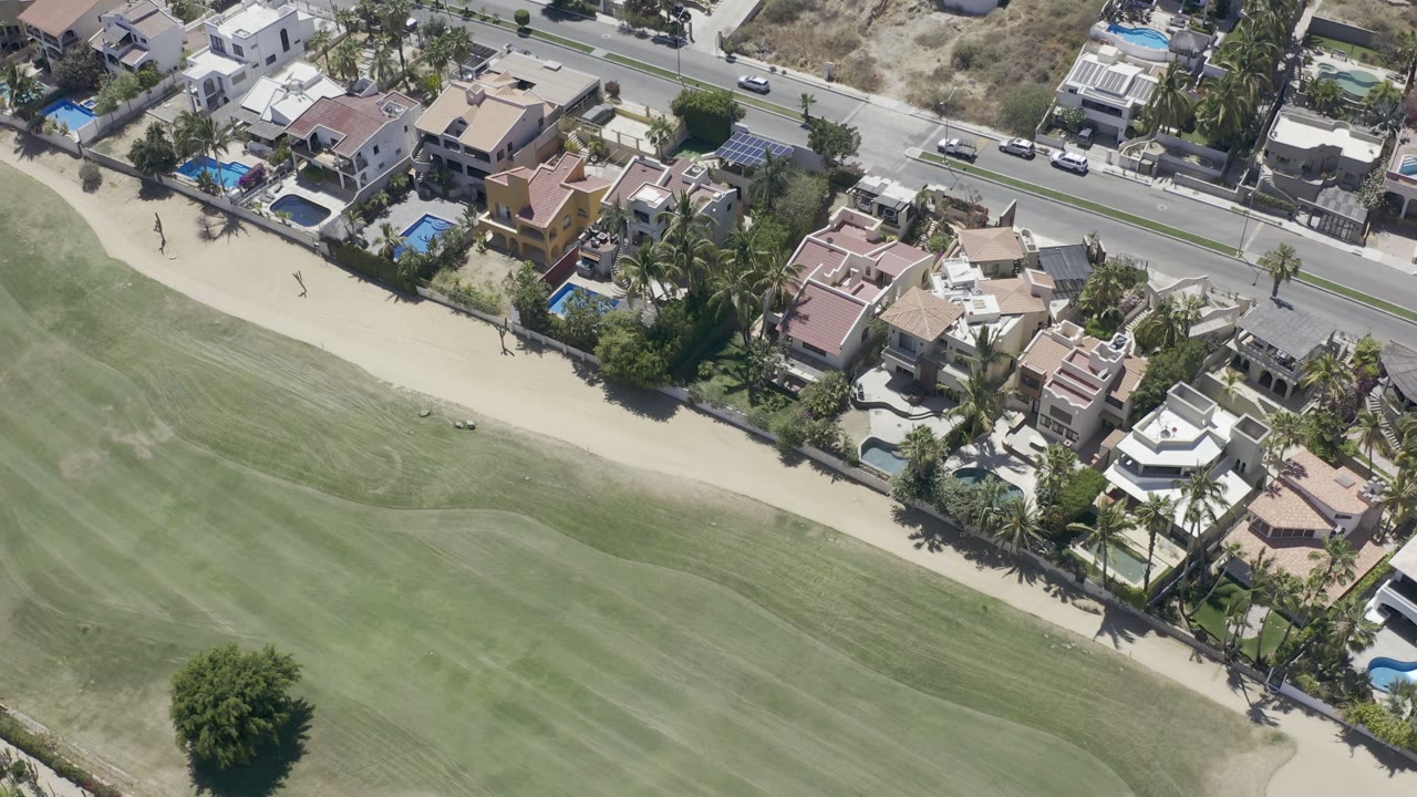 Aerial Los Cabos view with more fairway visible at the bottom, a row of homes and pools near the center, roadside cars above, dry terrain beyond, and no visible people.