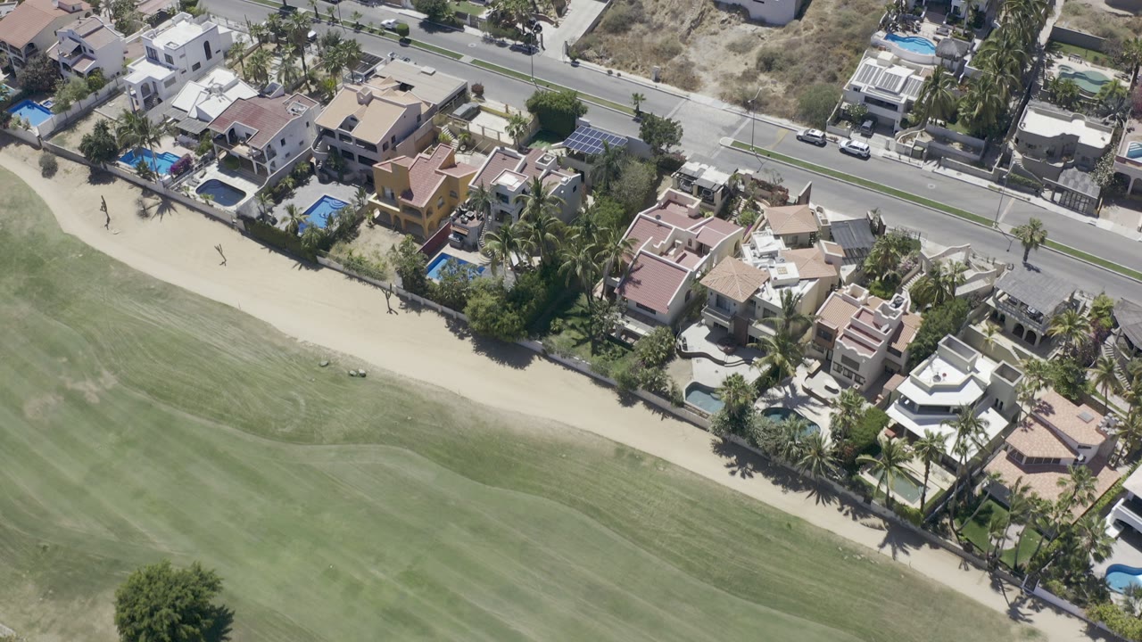 Overhead view of a green field beside homes, pools, palm trees, and a road.