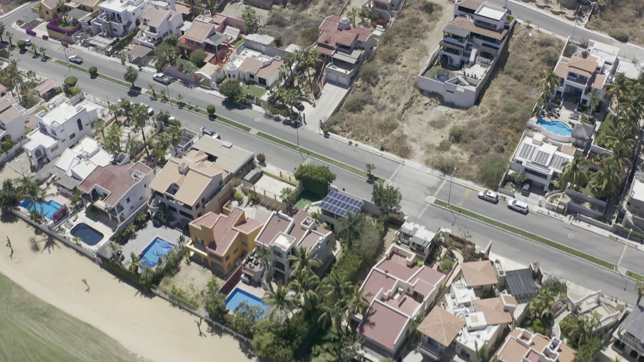 Aerial view of houses, road, pools, cars, and grass near a beach edge.