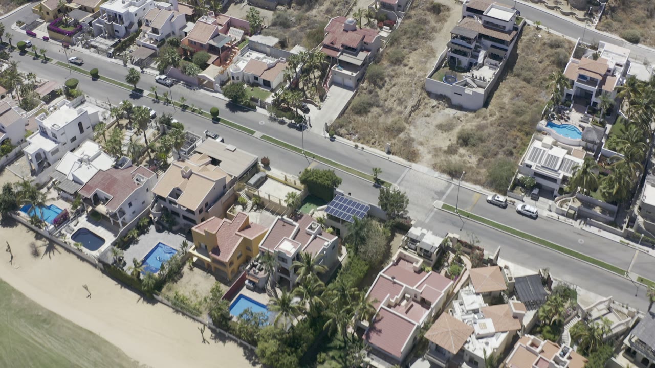 A slightly later aerial frame over Los Cabos residences shows the road running across the center, walled homes below it, dry lots above it, pools, palm trees, and tiny distant people near the lower left edge.