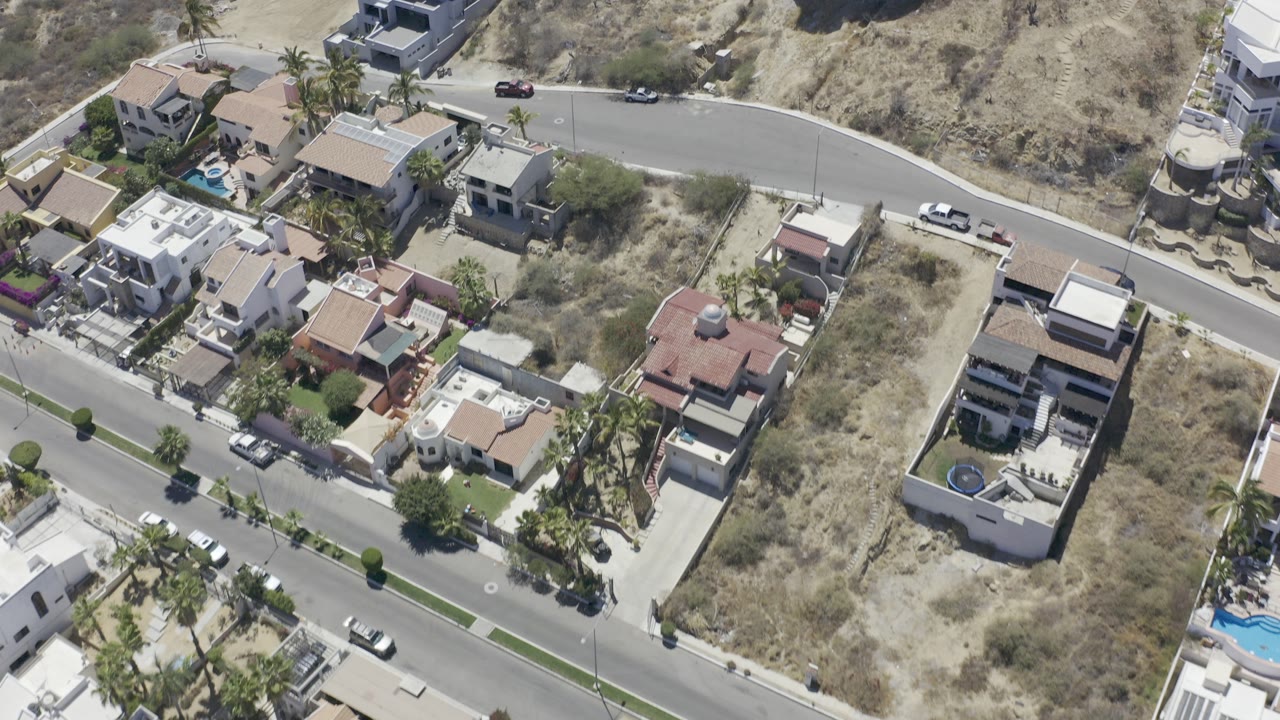 High aerial view over Los Cabos houses, a curving road, parked vehicles, palms, and dry hillside terrain, with no visible people.