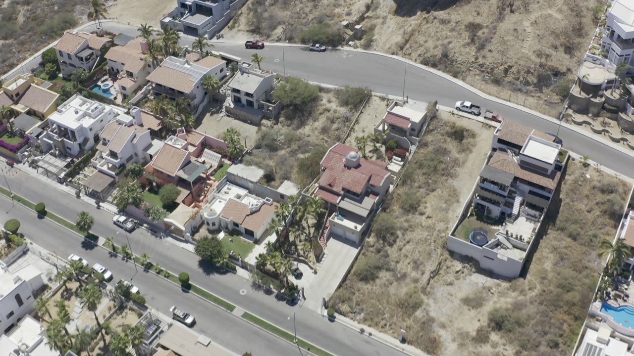 Aerial frame over Los Cabos residential roofs, small blue pools, palm trees, parked cars, and pale scrub-covered hillside, with no visible people.