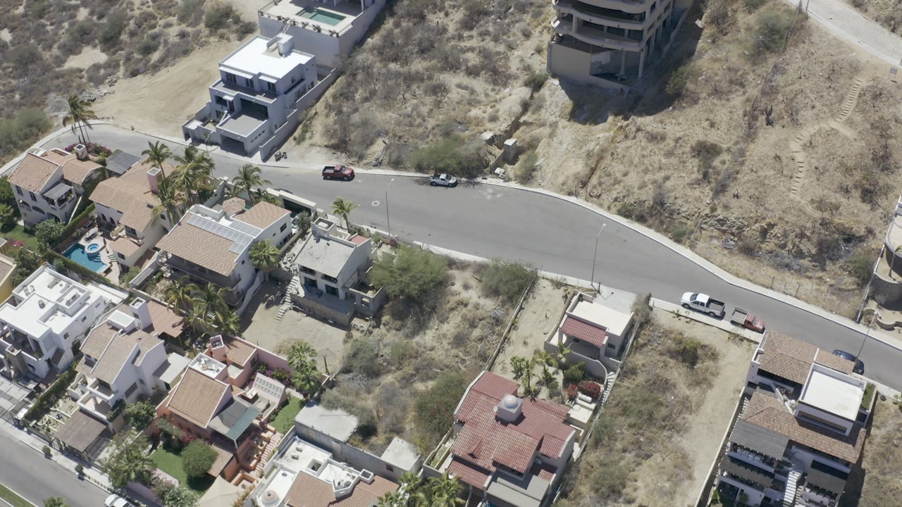 Aerial Los Cabos, Mexico view with tiled and flat-roof homes, palms, a small pool, parked cars along a curving street, and dry undeveloped land, with no visible people.