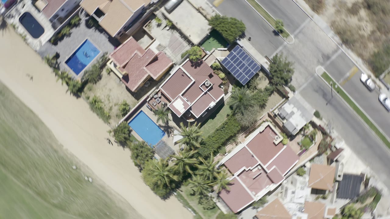 Overhead view of homes, pools, road, sand, and green coastal edge.