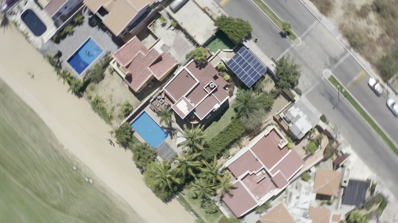 Overhead Los Cabos, Mexico view of residential lots with pools and palms, a paved road with small cars, sand, and green shoreline area, with no visible people.