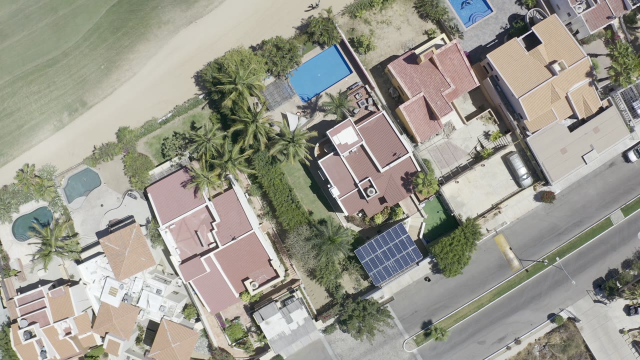 Aerial frame over a Los Cabos residential block, showing red tile roofs, palms, pools, a diagonal street, parked vehicles, and no discernible people.