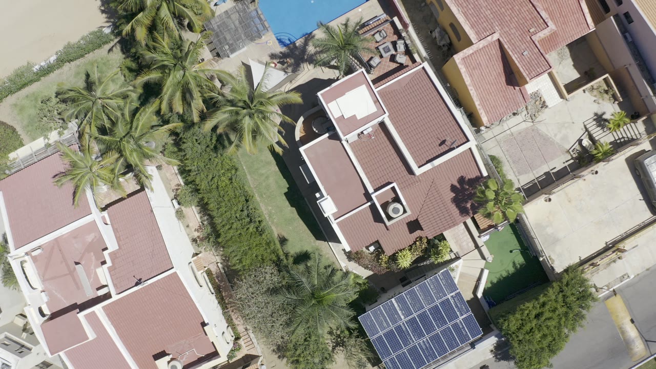 Second overhead moment in Los Cabos, Mexico, keeping the red-roofed house near center while palms, pale paving, hedges, pool water, and a solar-panel array remain visible with no visible people.