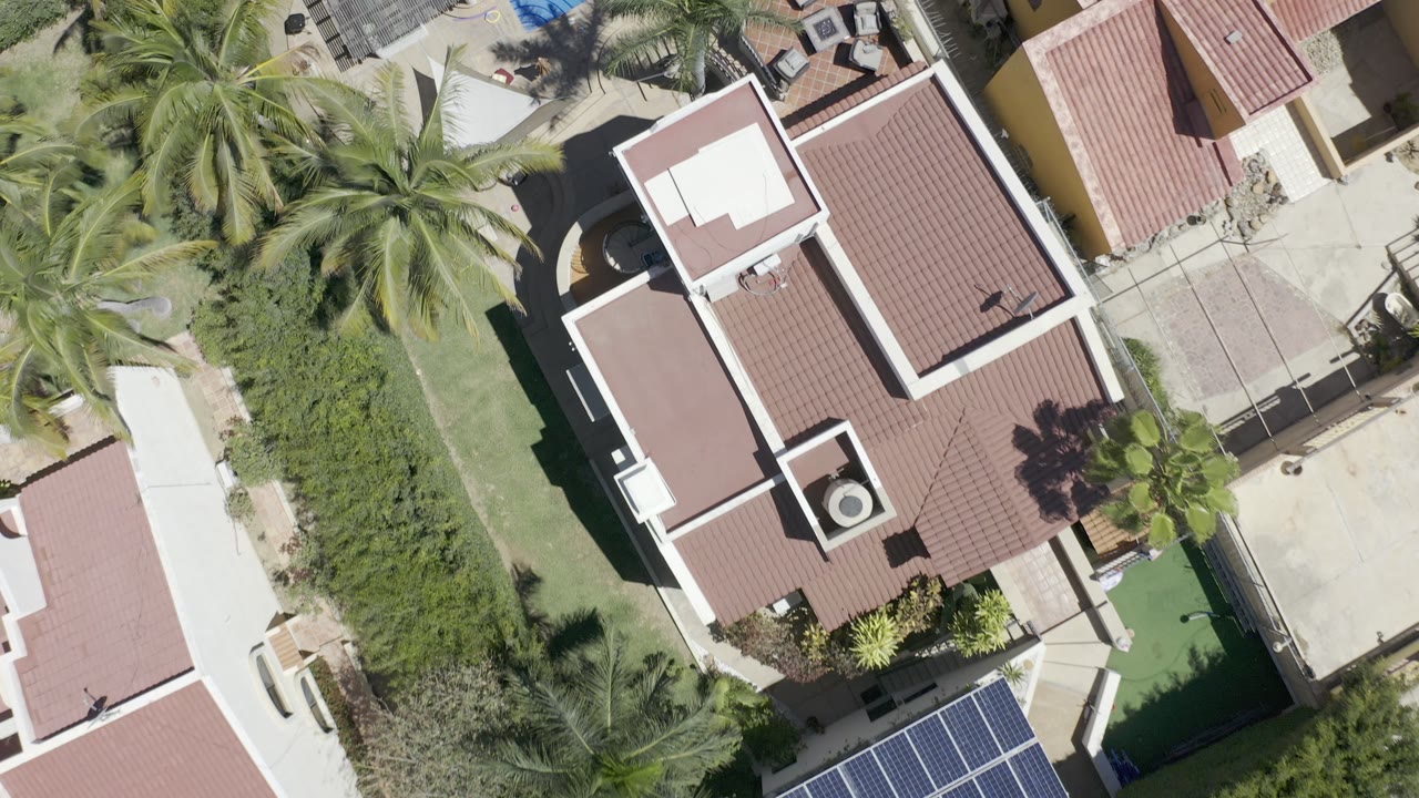 Top-down Los Cabos frame with a red tile roof centered beside palms, dense green hedges, pale paving, and no visible people.