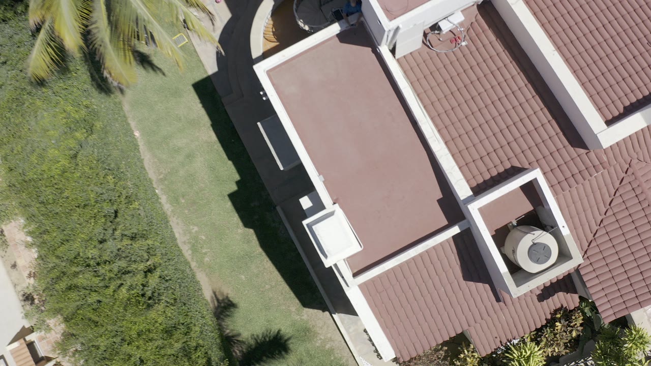 Top-down aerial frame of a Los Cabos villa roof with reddish tiles, white parapet walls, green lawn, shrubs, palm fronds, and no visible people.