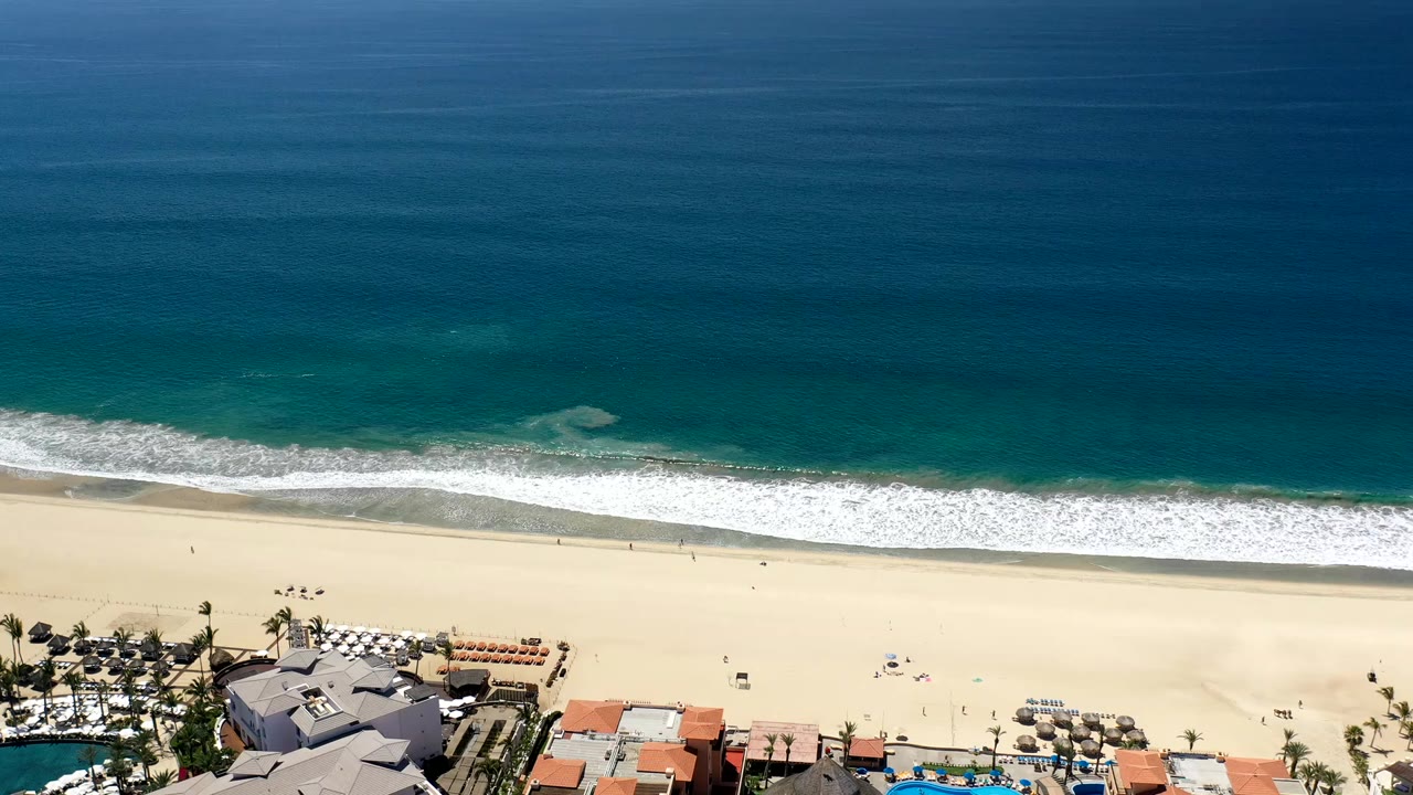 Aerial view of blue ocean, white surf, sandy beach, and beachfront rooftops.