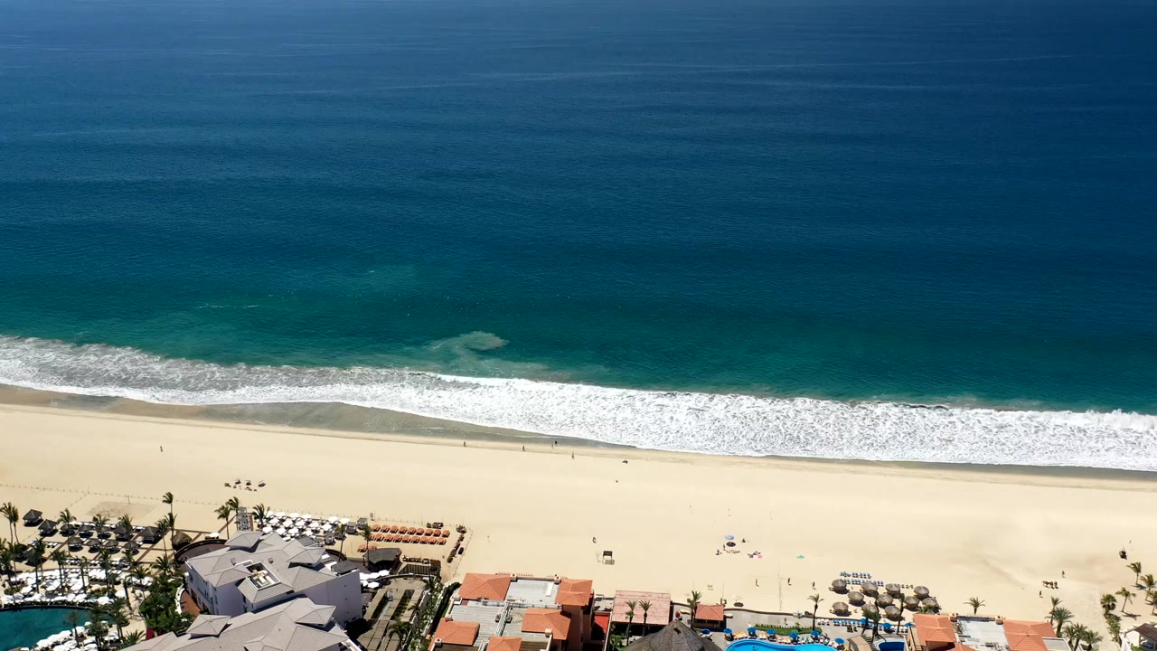 A Los Cabos aerial view shows a broad band of blue water, a fresh line of white surf, pale sand, small beach figures, umbrellas, palms, and cropped rooftops below.