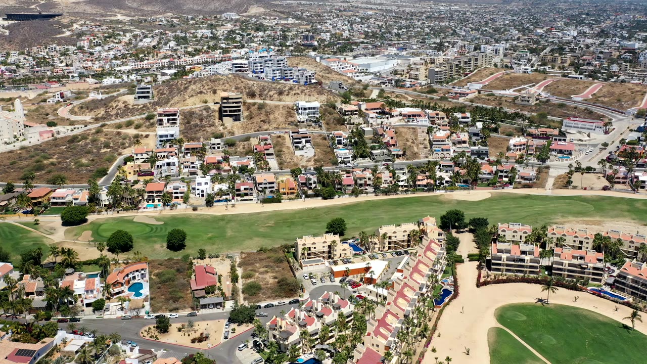 Opening aerial frame over Los Cabos, Mexico, with green fairways below, residential buildings on dry hillsides, dense development beyond, and no visible people.