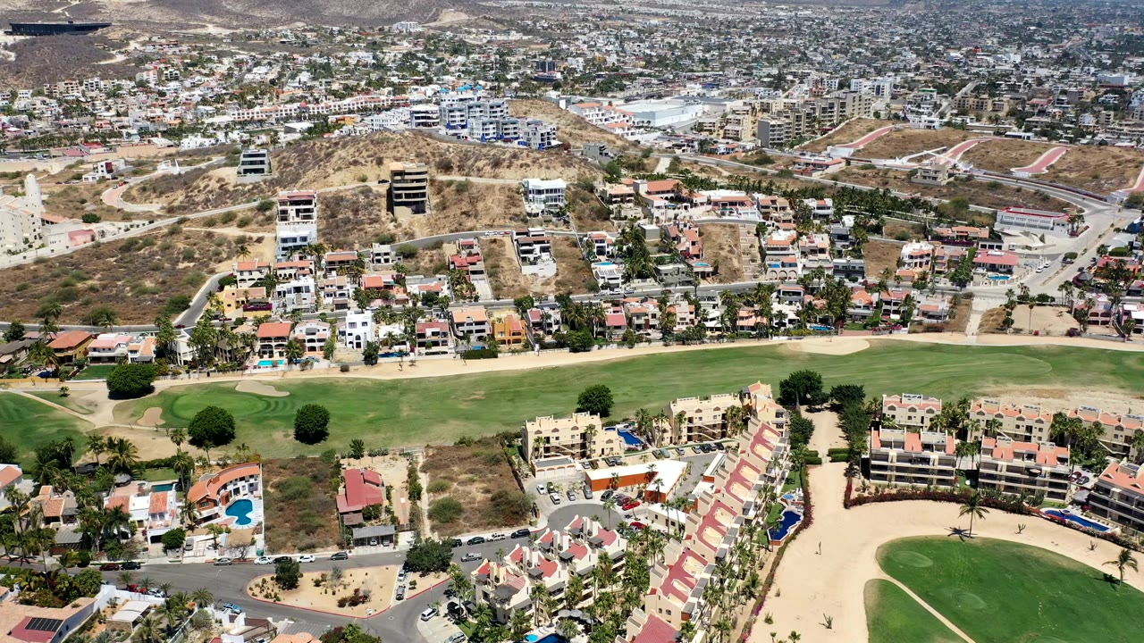Los Cabos aerial frame with a broad golf fairway in front, clustered homes and condos across the middle, dry hills behind them, and no visible people.