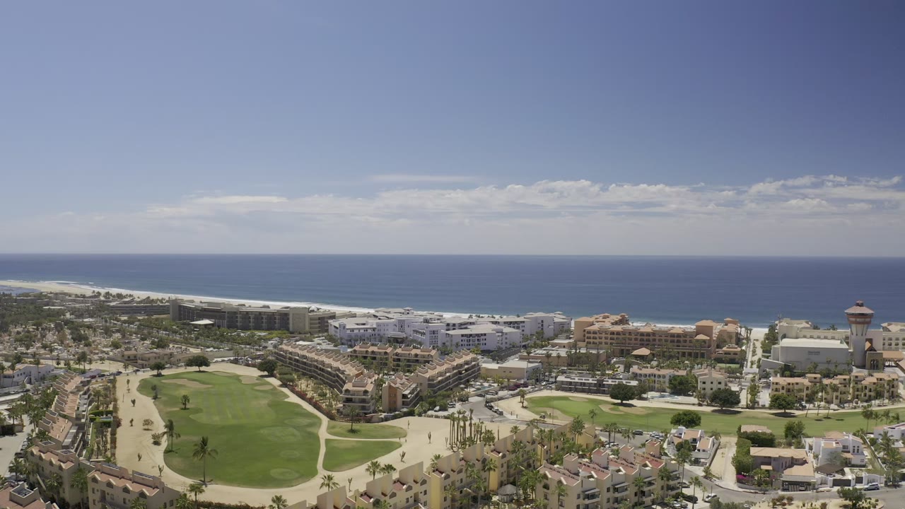 Wide aerial frame over Los Cabos, Mexico, showing green open space, resort-style buildings, palm-lined roads, a pale beach, and the blue ocean horizon with no clearly visible people.