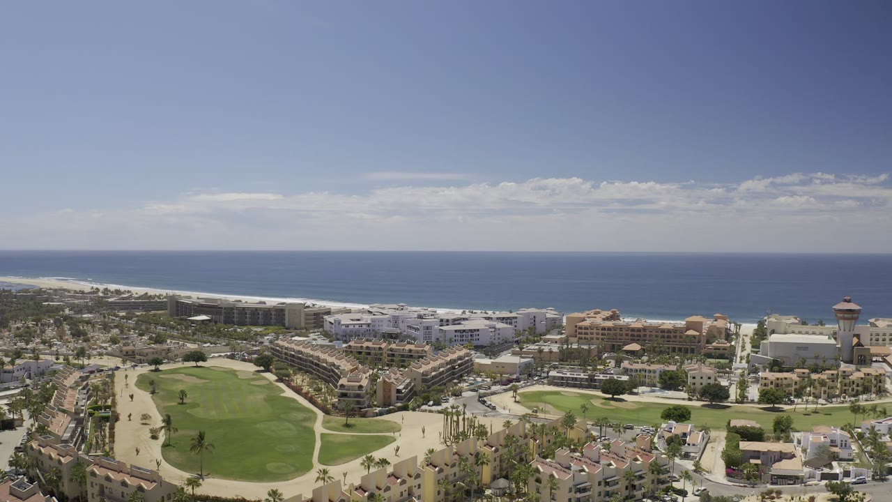 Elevated Los Cabos view with the ocean stretching across the background, light-colored beachfront buildings in the midground, palm trees along roads, and a large green lawn in front with no clearly visible people.