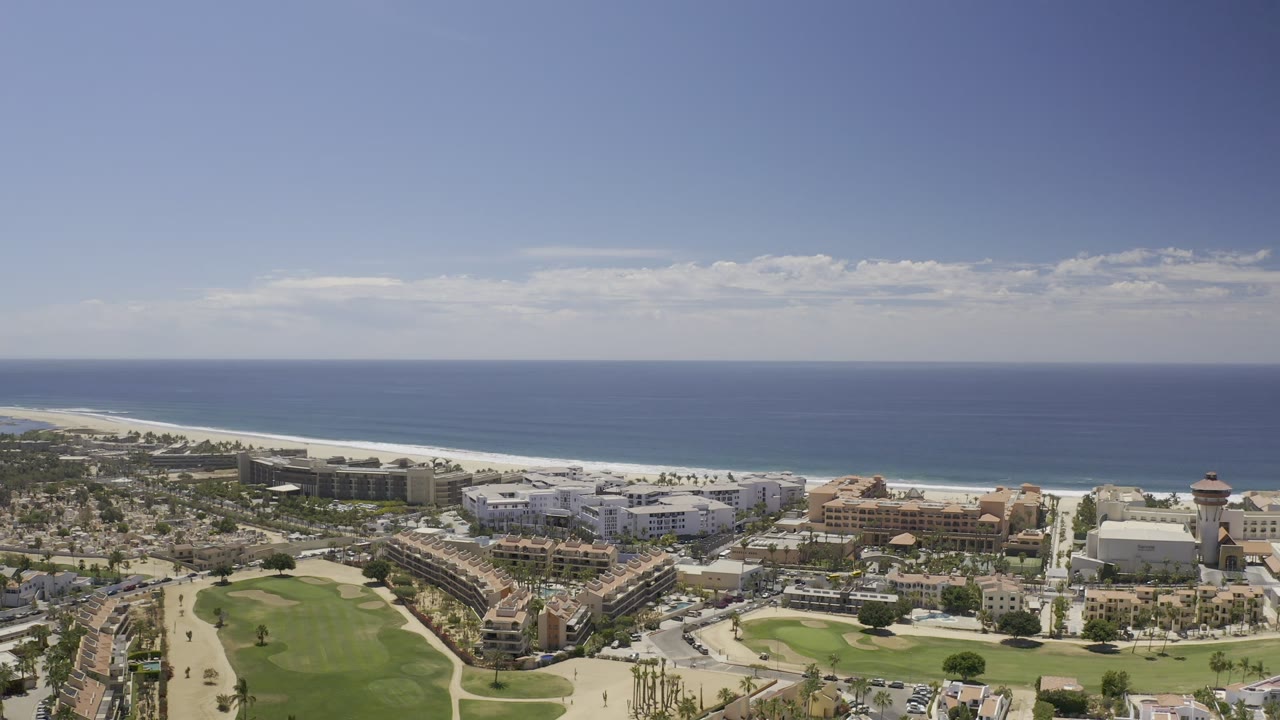 Aerial Los Cabos frame showing hotel blocks, landscaped greens, roads, a long sand beach, and open ocean under scattered clouds, with no clearly visible people.